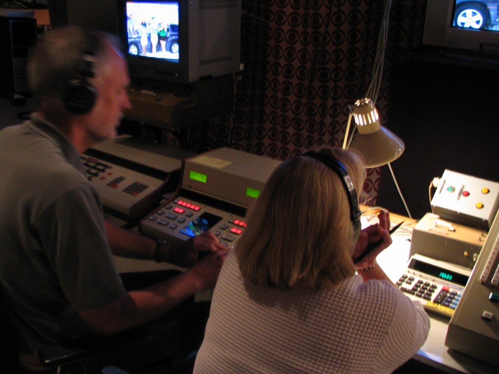 Two individuals in a control room during a television production, with equipment in front of them and a monitor showing a scene from the show.
