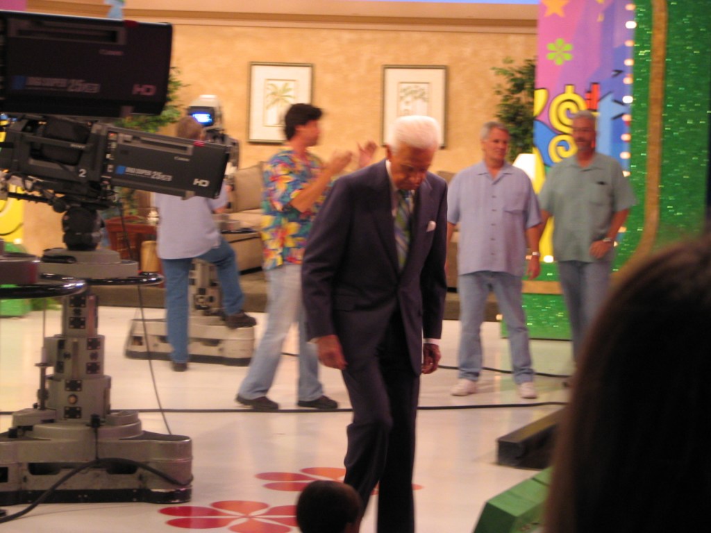 Bob Barker walking on stage in a television studio, with crew members and cameras in the background.