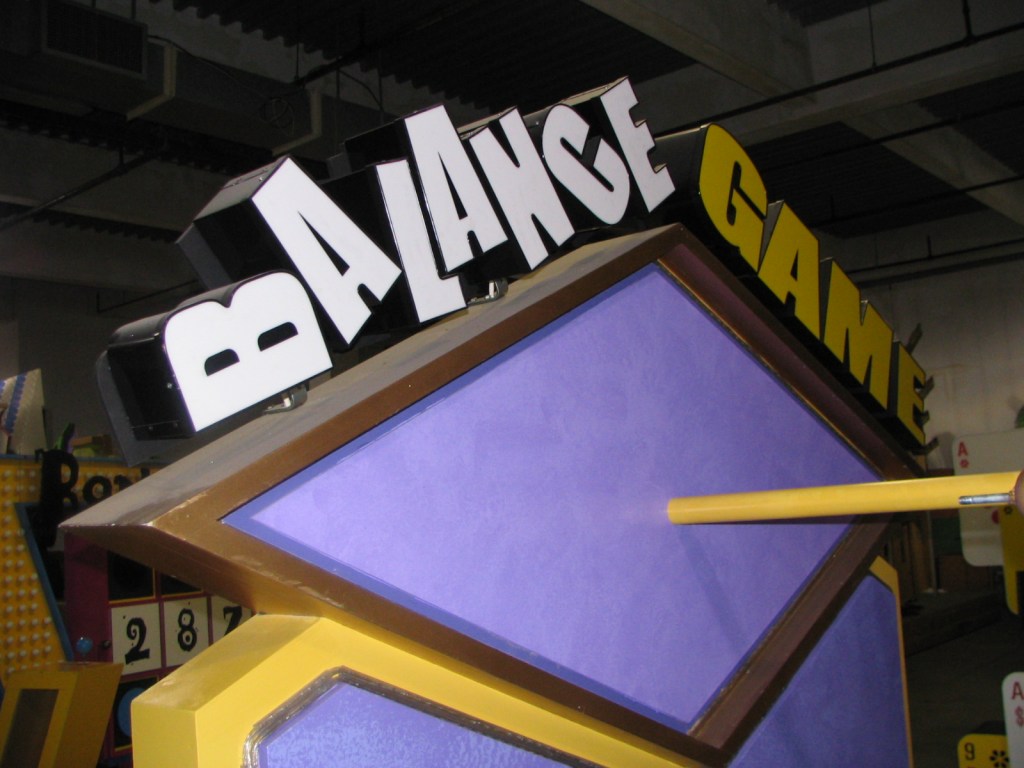 Close-up of a sign featuring the words 'BALANCE GAME' in bold letters against a colorful backdrop, taken backstage at a TV game show.