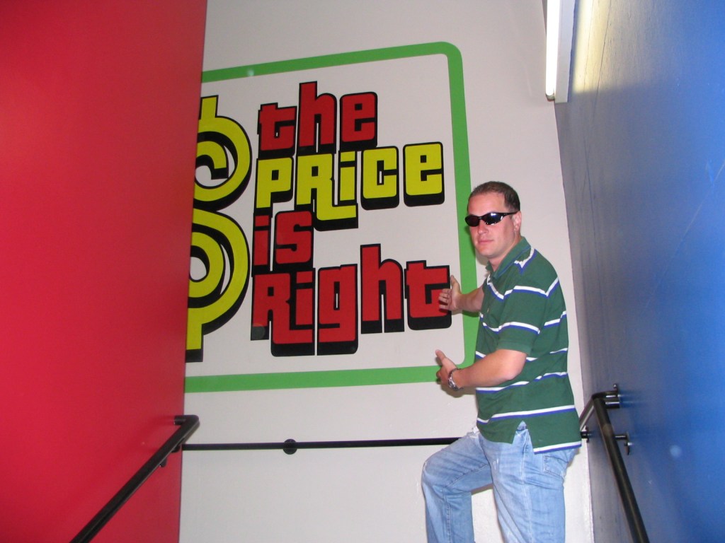 A person wearing sunglasses poses next to a large colorful sign that reads 'the Price is Right' in a hallway.