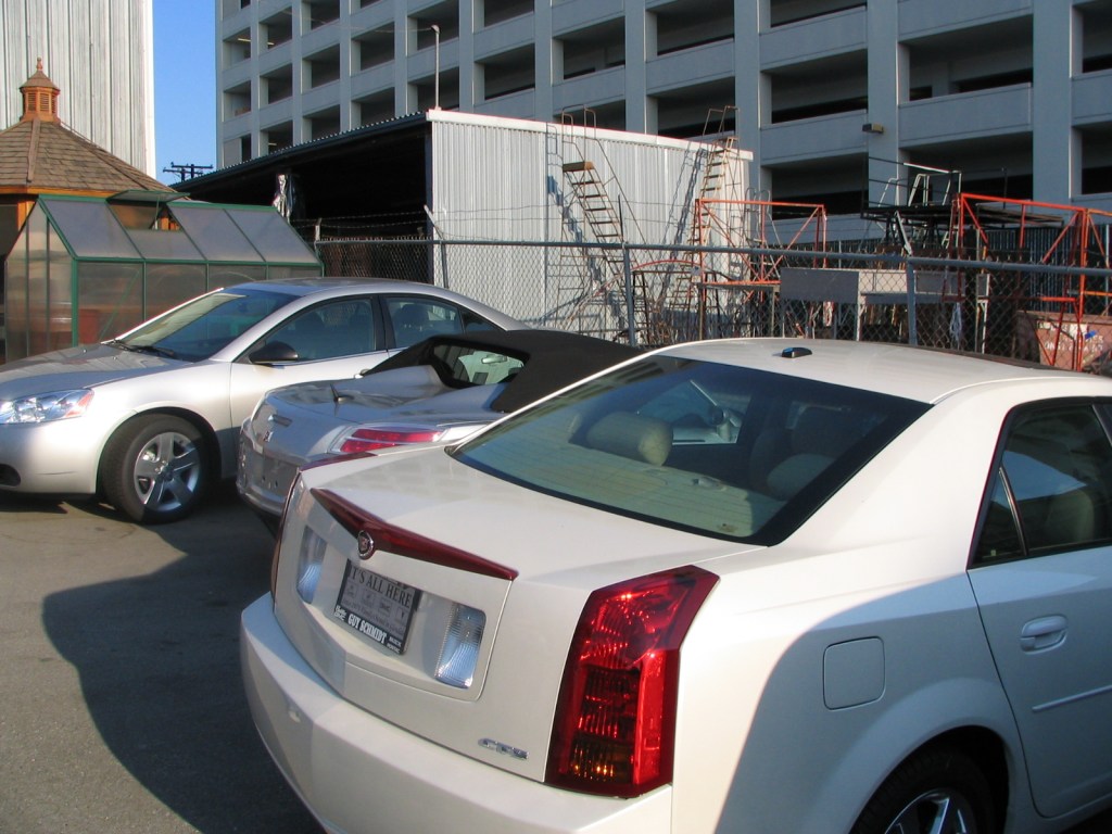 Parking area with multiple cars, including a silver sedan and a convertible, near a building with a fence and construction equipment in the background.