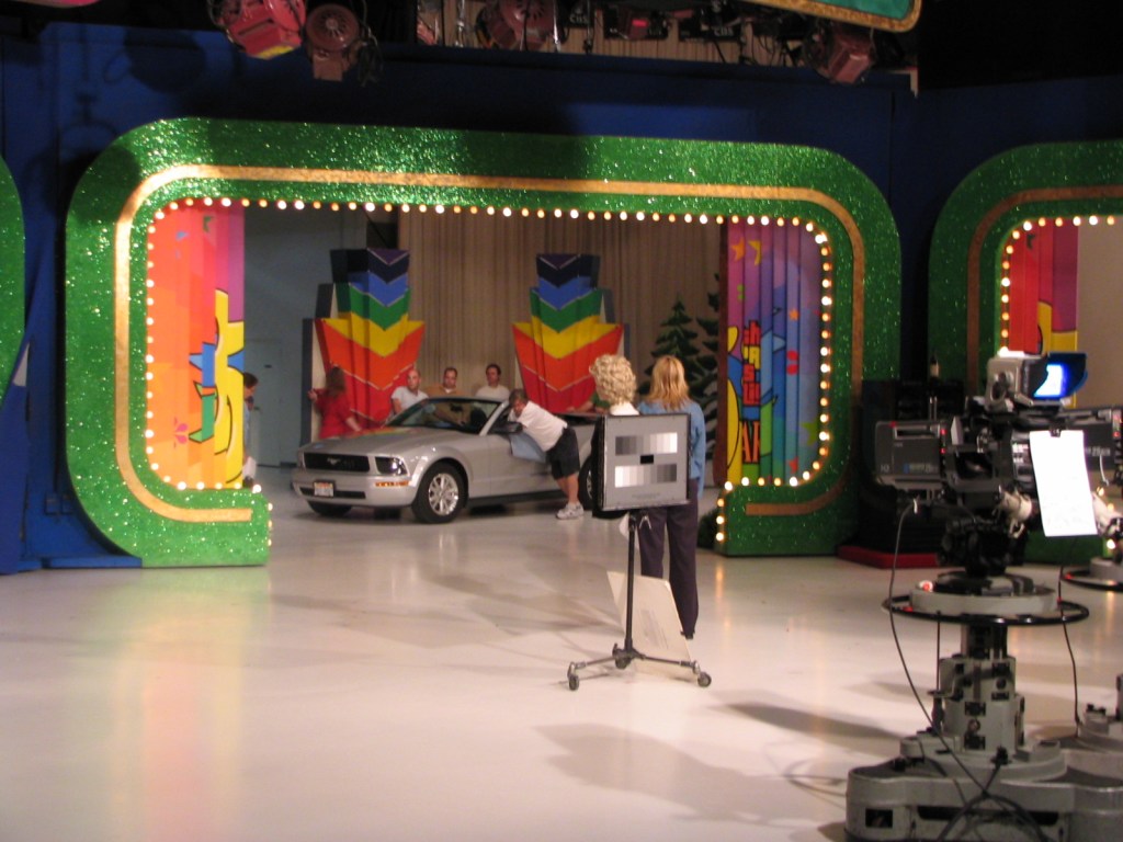Backstage view of a television studio during the taping of a game show, featuring a silver car and colorful set designs with bright lights.
