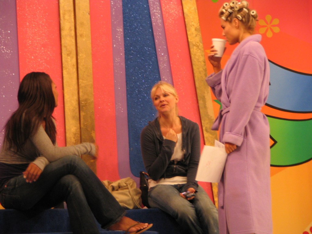 Three women backstage at a show, one wearing a purple robe with curlers in her hair, while the others are sitting and talking.