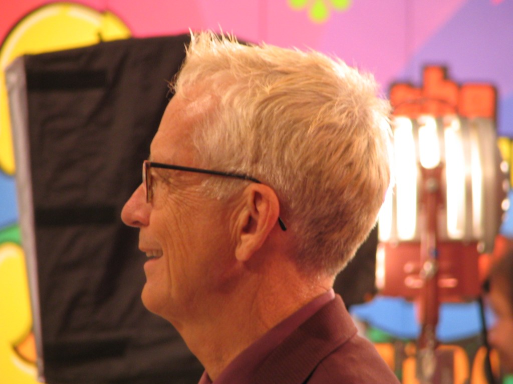 A side profile of a man with short, light-colored hair and glasses, smiling in a colorful background with studio lighting.