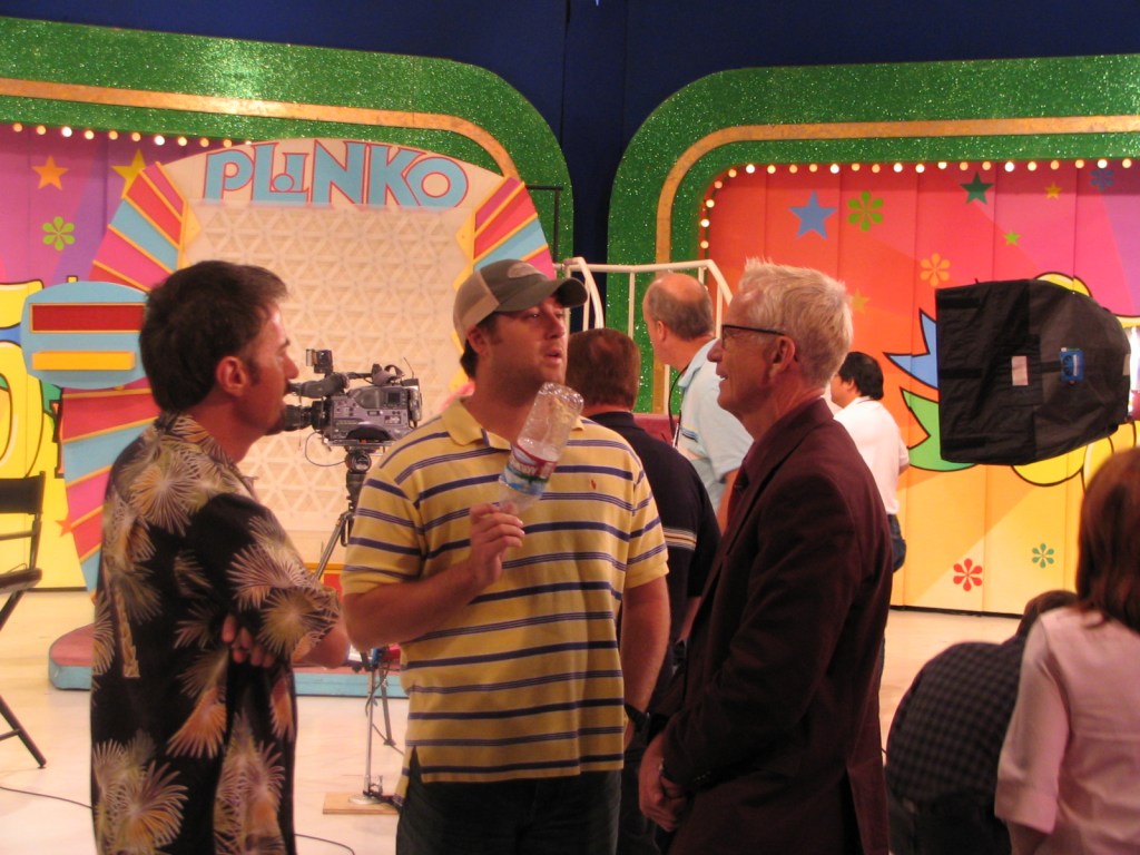 Backstage scene at the Plinko game set with crew members discussing preparations, colorful set decorations in the background.