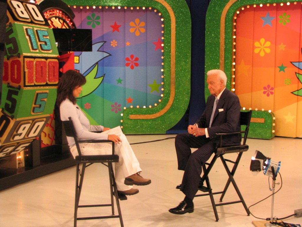 A televised interview with a woman sitting on a chair, engaged in conversation with Bob Barker in a striped suit, set against a colorful game show backdrop.