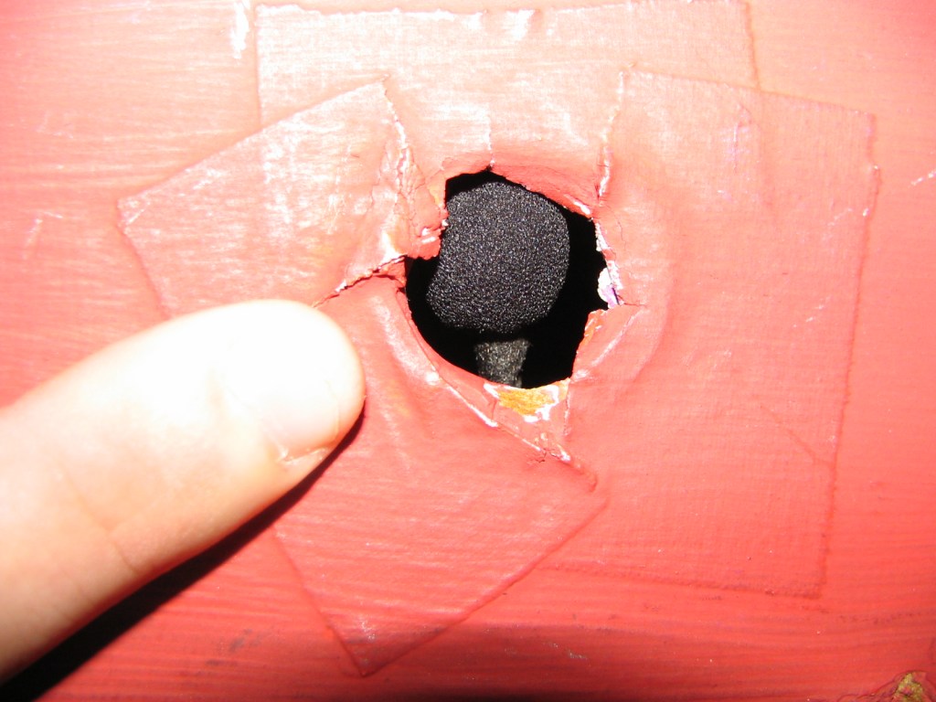 A close-up of a finger pointing at a hole in a red surface, revealing what appears to be a microphone or sound equipment inside.