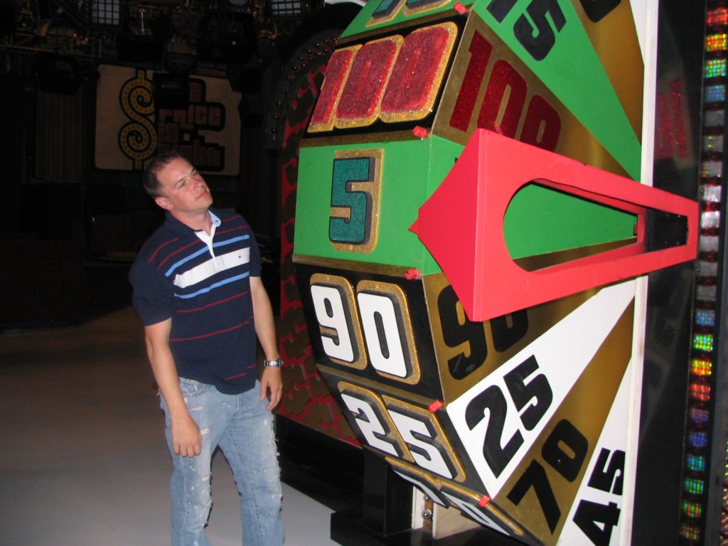 A man stands in front of a colorful game show wheel, looking thoughtfully at the numbers on it, with the set's vibrant backdrop visible.