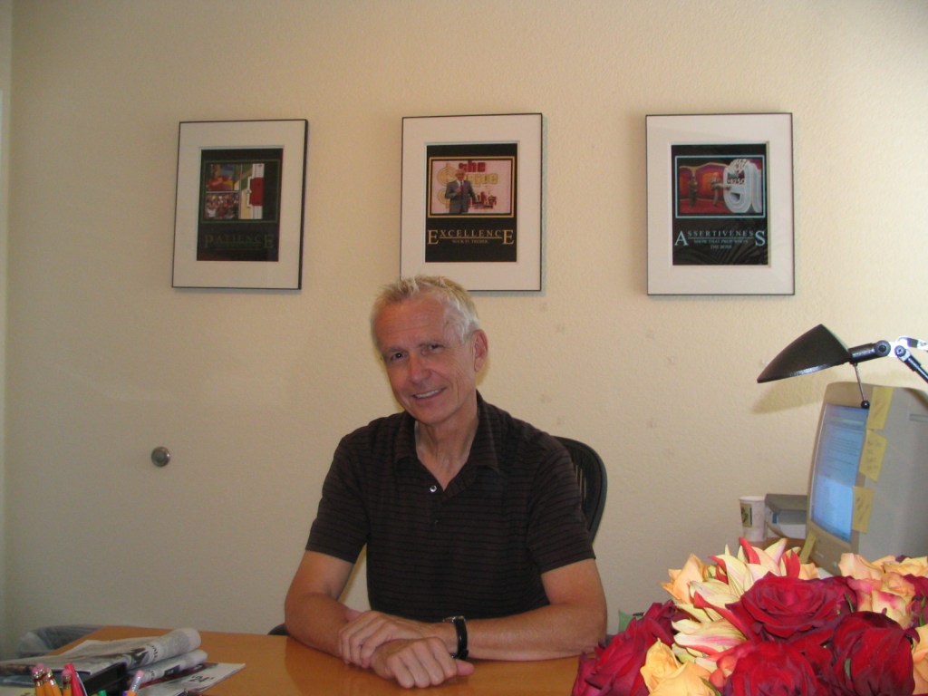 A man sitting at a desk with a friendly smile, surrounded by framed motivational posters on the wall and a bouquet of red roses on the table.