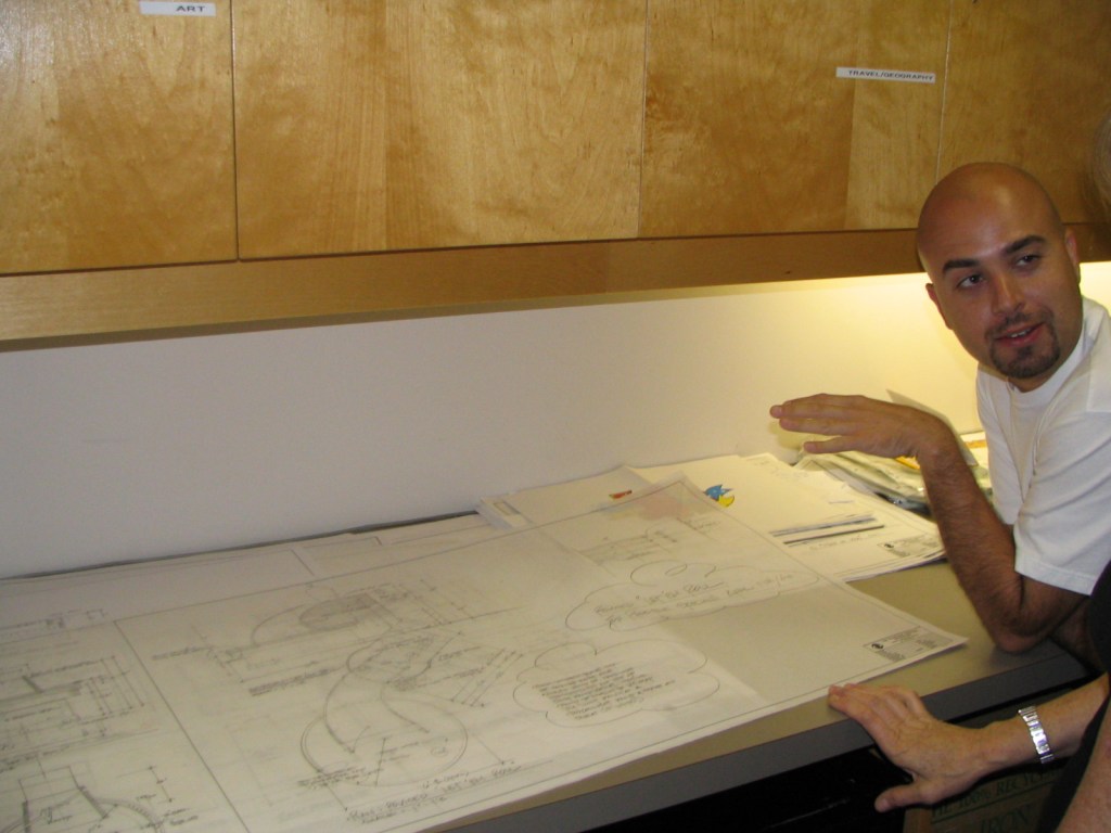 A man discussing design plans at a table covered with architectural drawings and sketches, with wooden cabinets in the background.