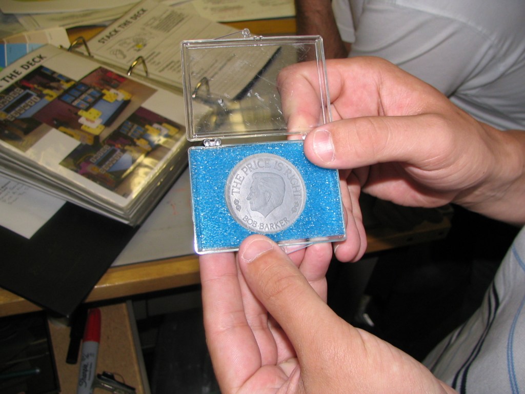 A person holding a commemorative coin in a transparent case with blue foam, featuring the inscription 'THE PRICE IS RIGHT BOB BARKER.'