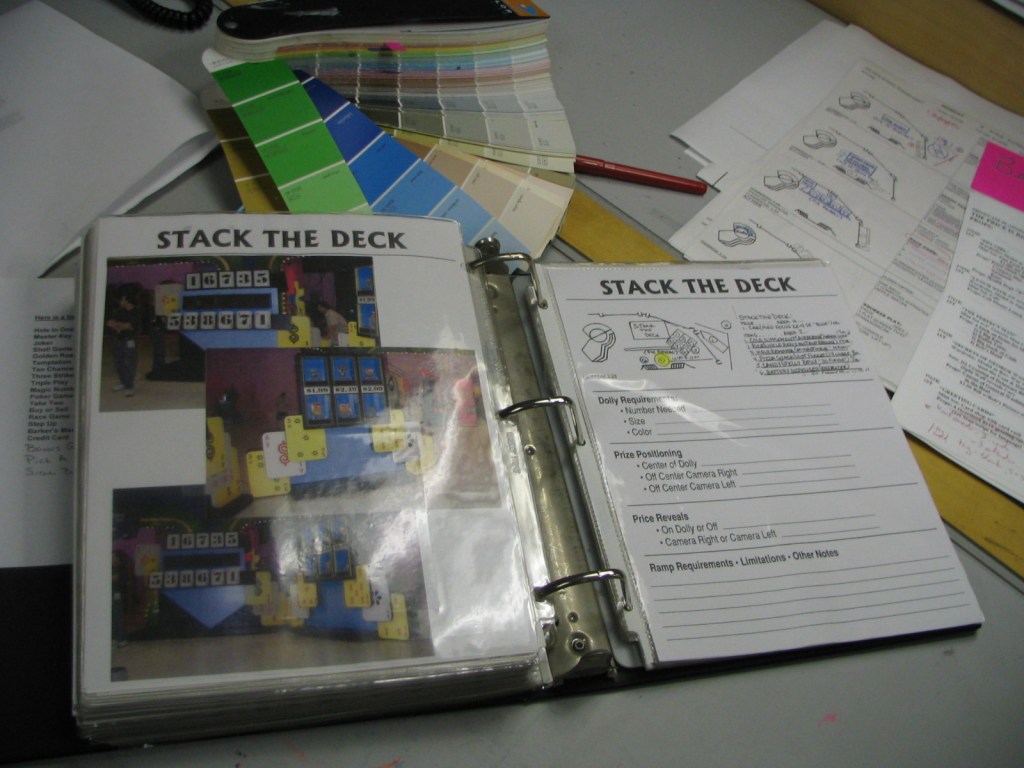 A binder opened to a page labeled 'STACK THE DECK,' containing images and notes related to the game setup for a television show. The background includes color swatches and scripts.