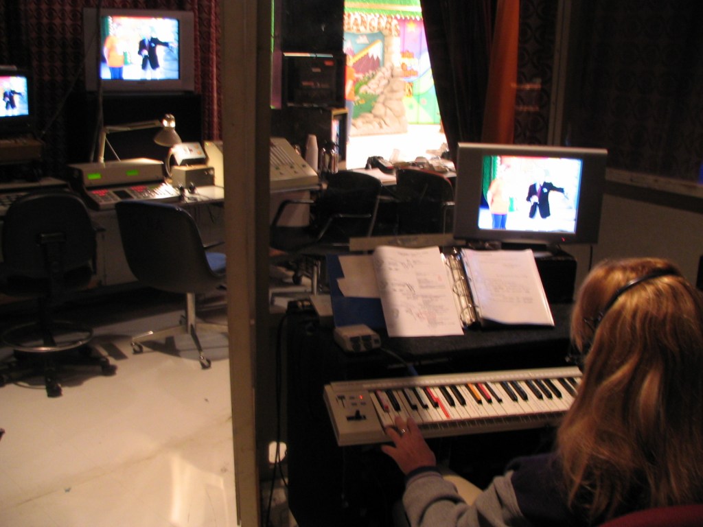 A backstage view of a television production room with a person operating a keyboard and monitoring screens showing a live broadcast.