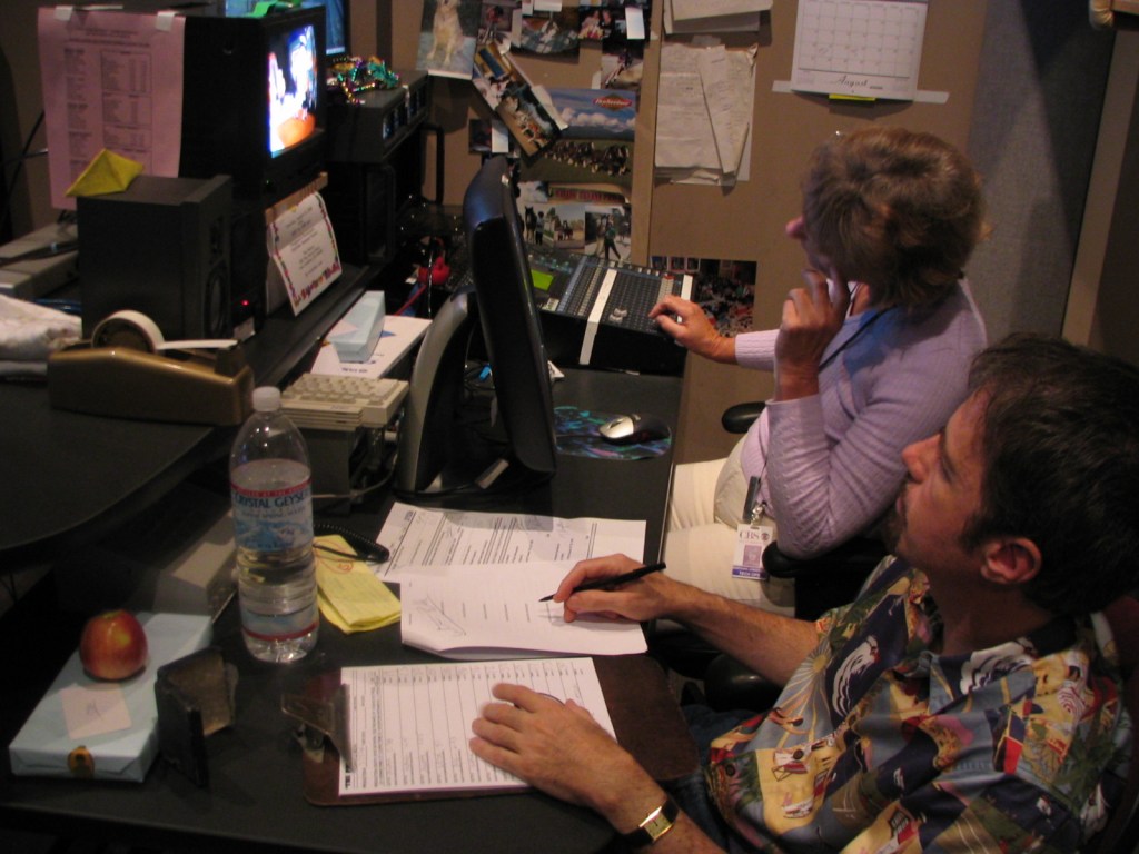 Two individuals working at a control station during a television show taping, surrounded by screens, paperwork, and office supplies.