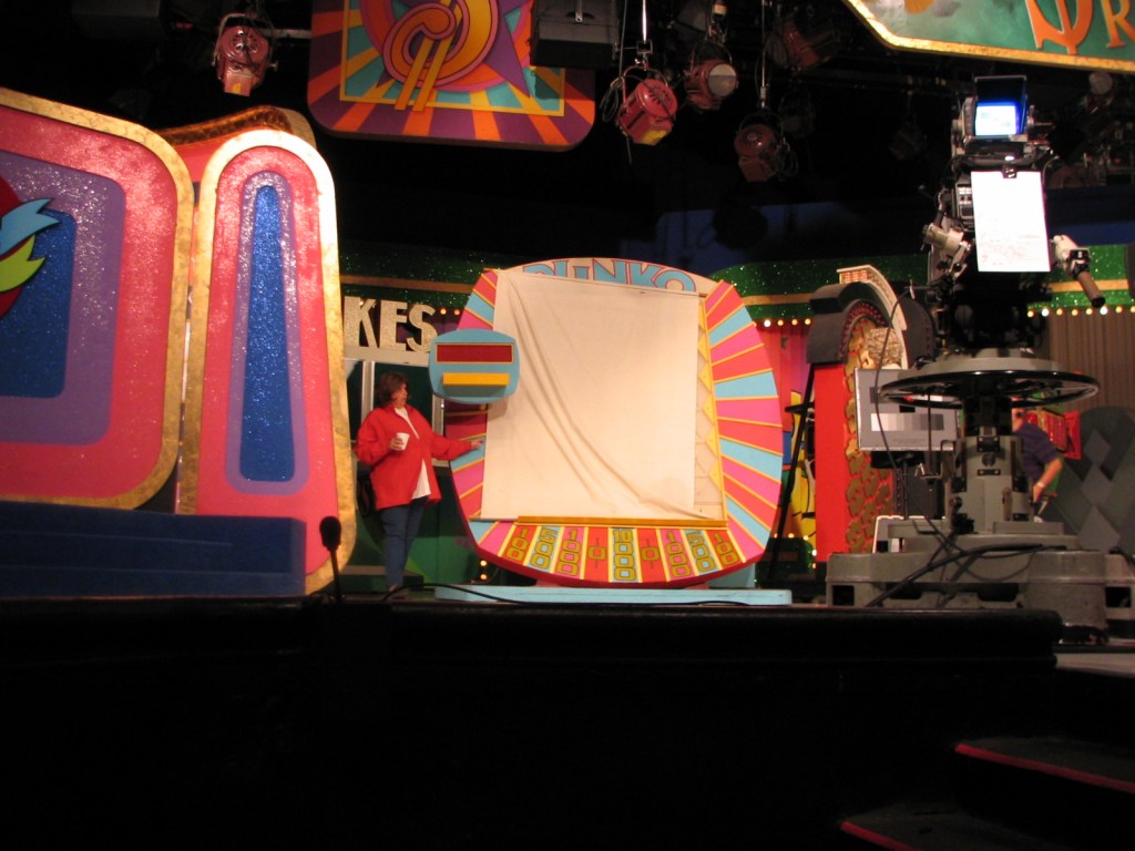 Backstage view of a television set during the taping of a game show, featuring a colorful game wheel and a crew member preparing for the next segment.