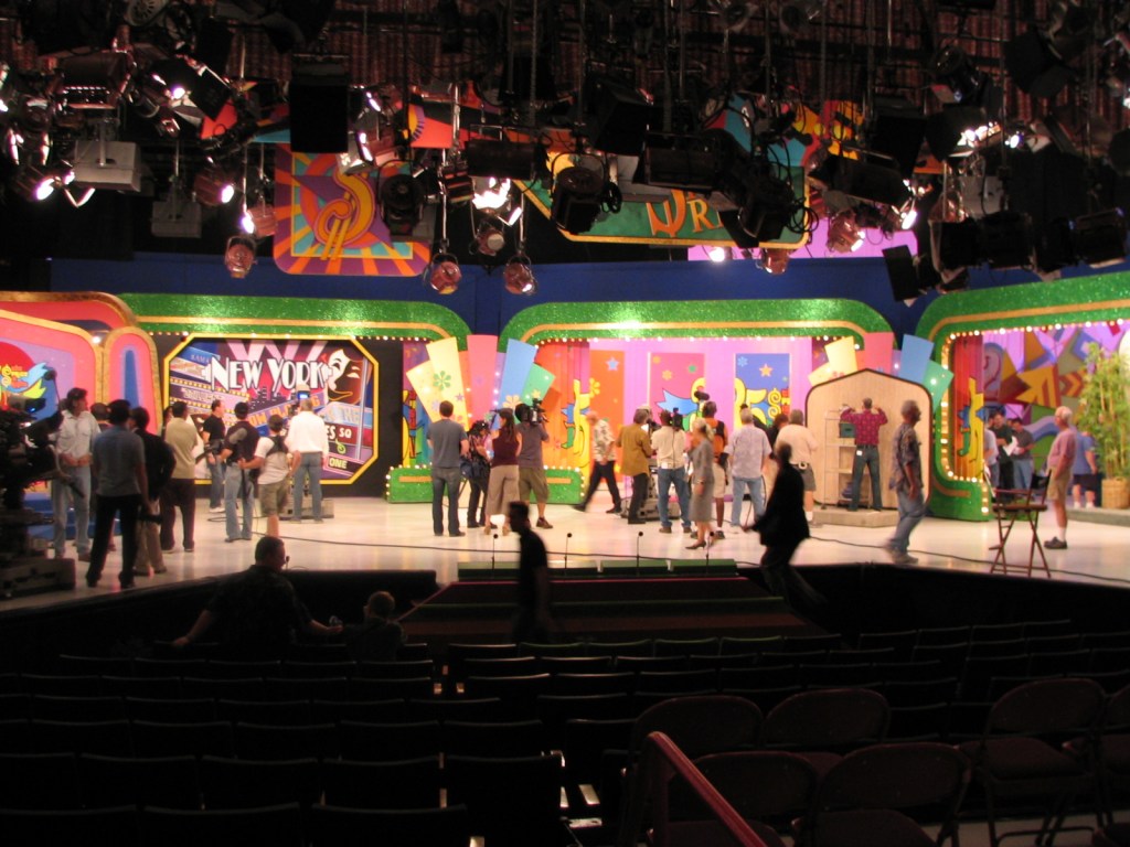 Backstage view of a TV set during a taping, with crew members and participants preparing for a show. Colorful set design visible in the background.