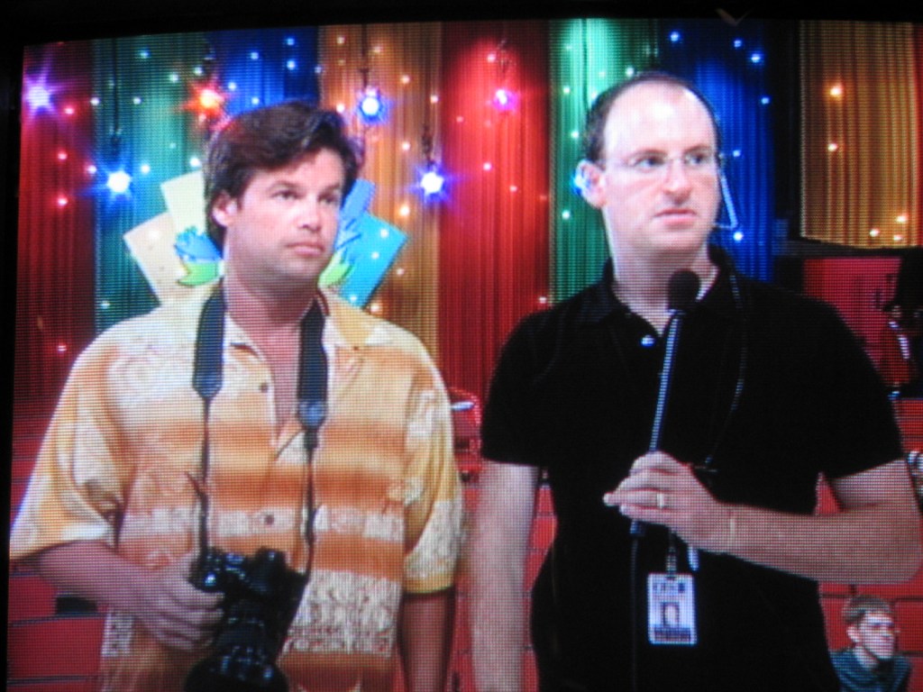 Two men standing backstage, one holding a camera and the other holding a microphone, with a colorful backdrop of lights and decorations.