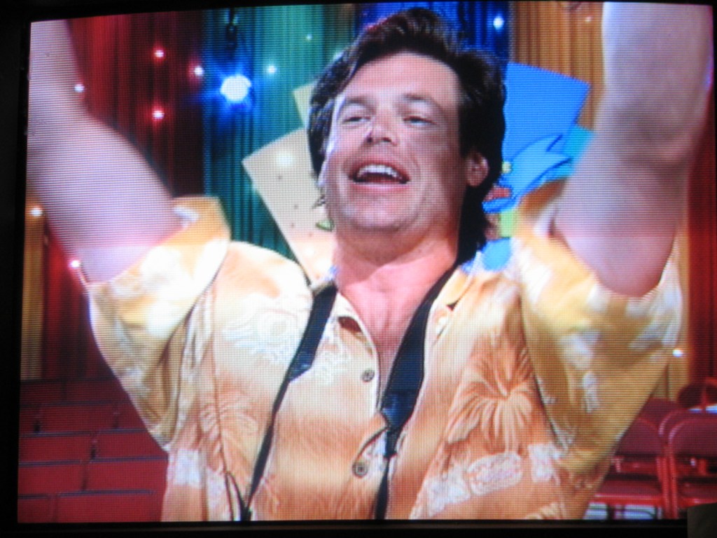 A man in a floral shirt smiling and raising his arms in excitement, with colorful stage curtains in the background.