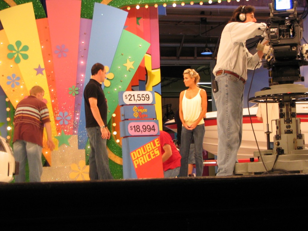 A behind-the-scenes photo from the set of The Price is Right, showing two crew members adjusting the game display with price tags. One woman stands nearby, waiting, while the colorful set background features bright decorations.