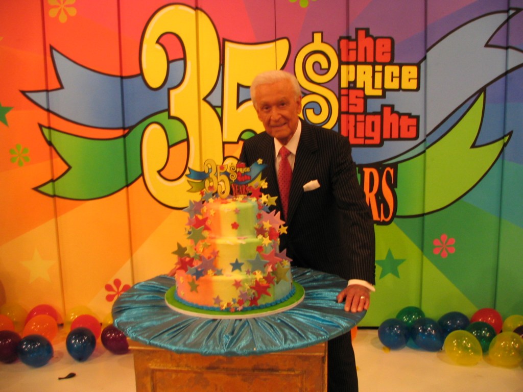 A man in a formal suit stands beside a colorful birthday cake celebrating 'The Price is Right's 35th anniversary, with festive decorations and a bright backdrop.