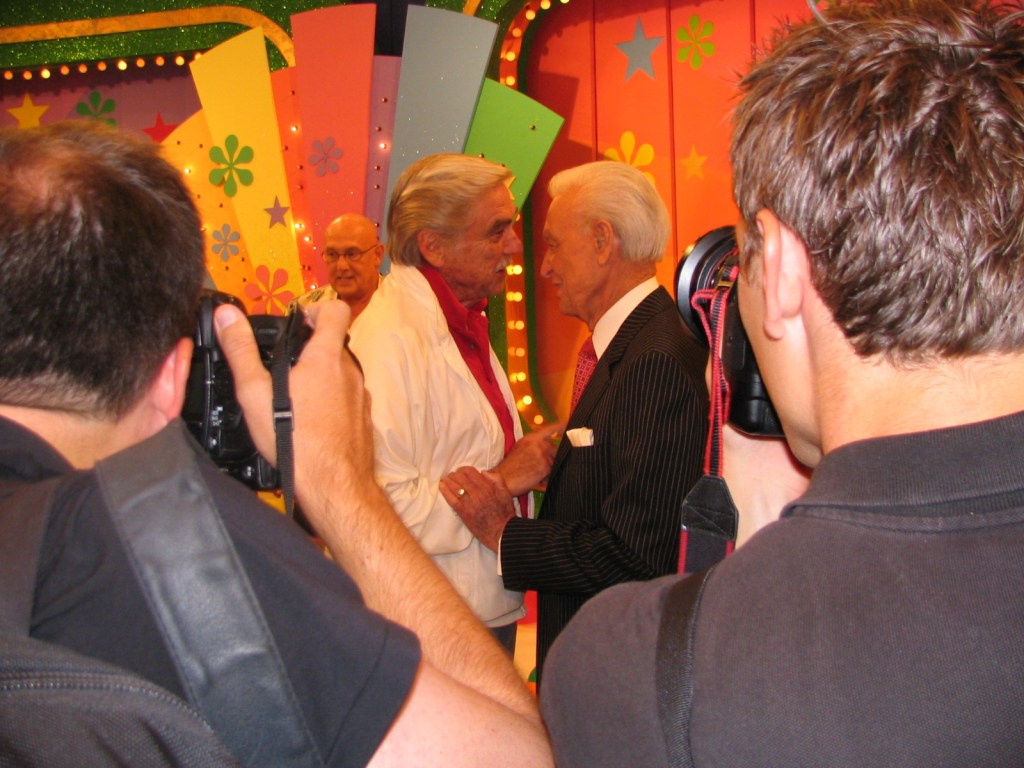Two men shaking hands on a colorful television set background, with photographers capturing the moment.