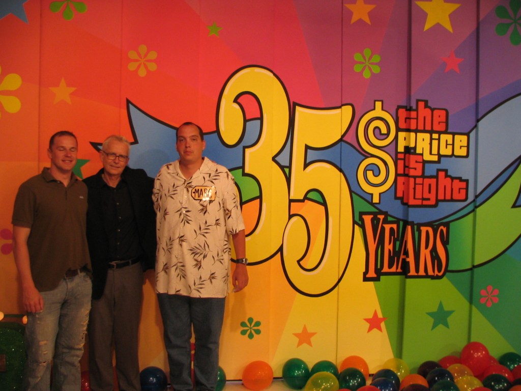 Two men pose alongside an older man in front of a colorful backdrop celebrating the 35th anniversary of 'The Price is Right'.