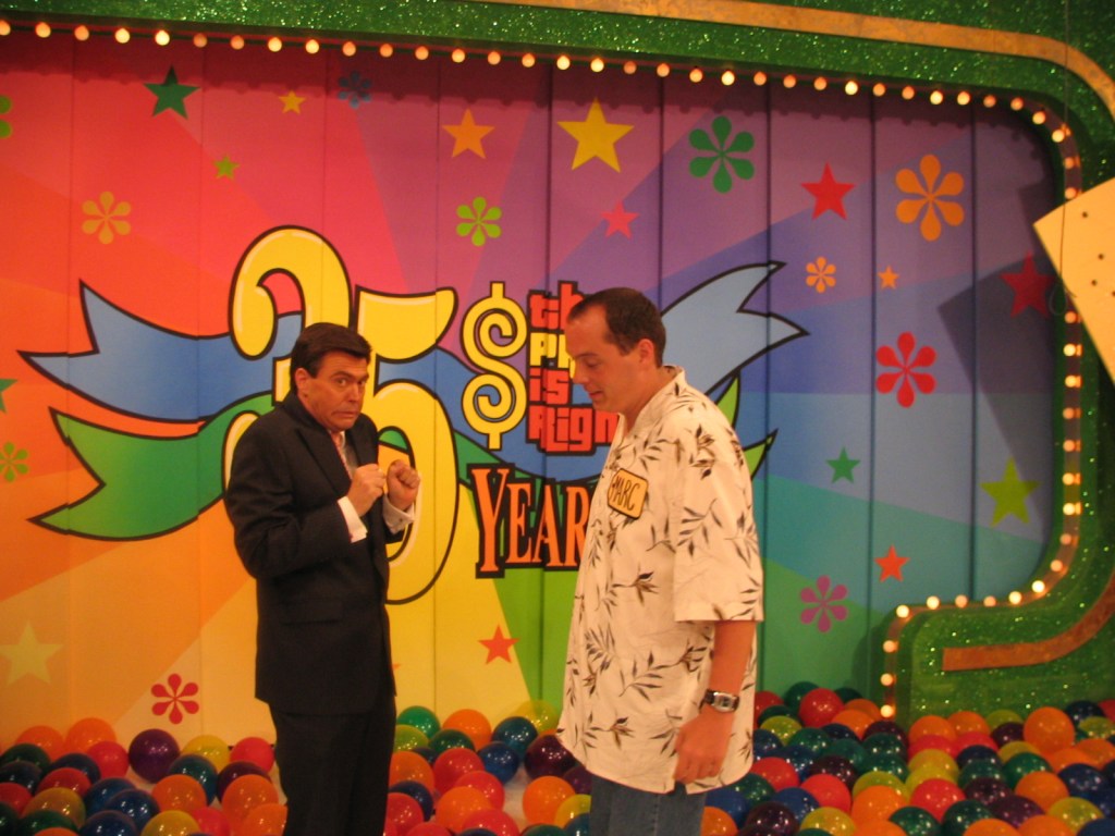 Two men in suits on stage celebrating the 35th anniversary of a game show, surrounded by colorful balls and a vibrant background.