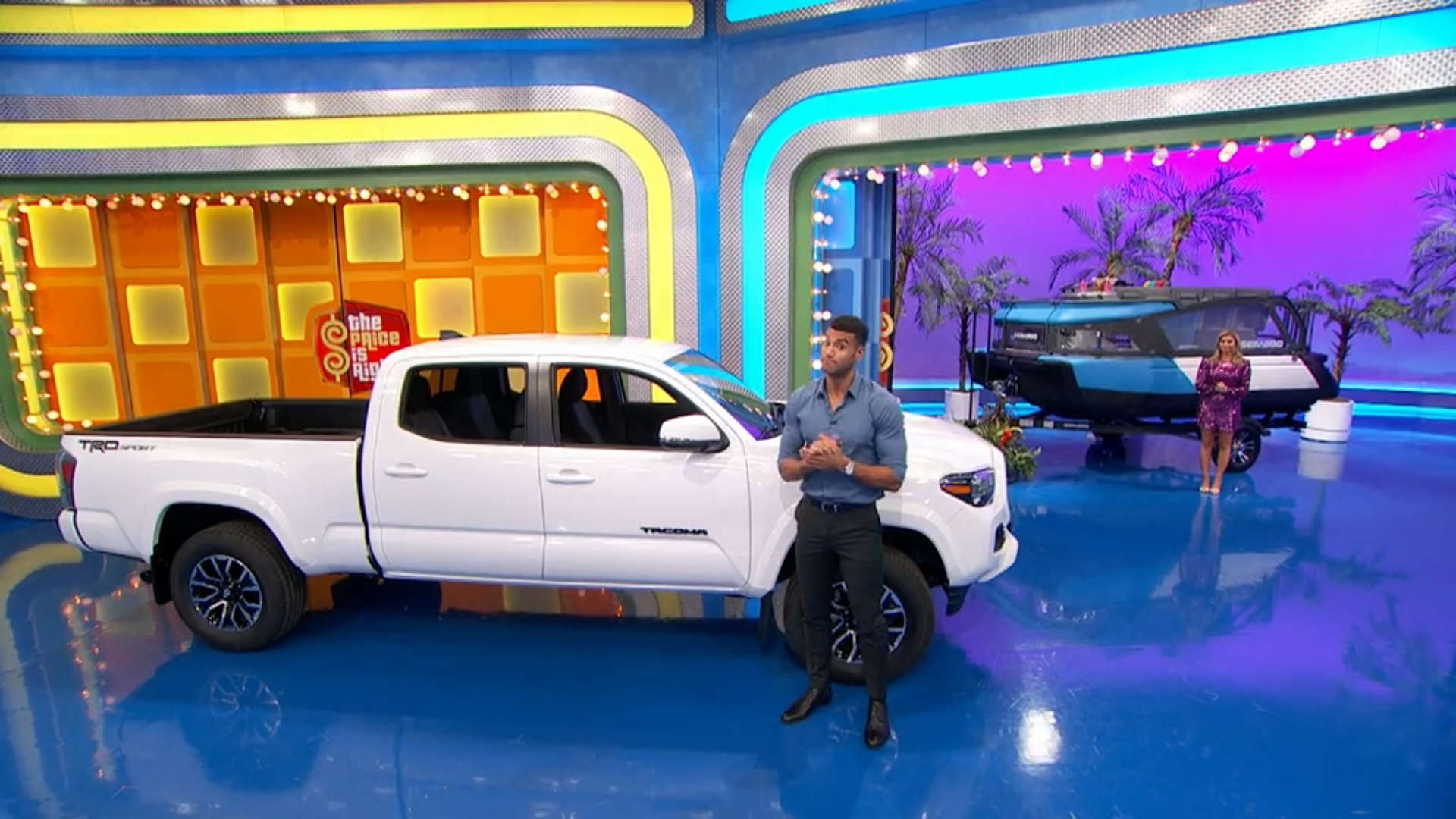 A contestant stands in front of a white pickup truck and a trailer, ready to play a game on a brightly colored TV set.