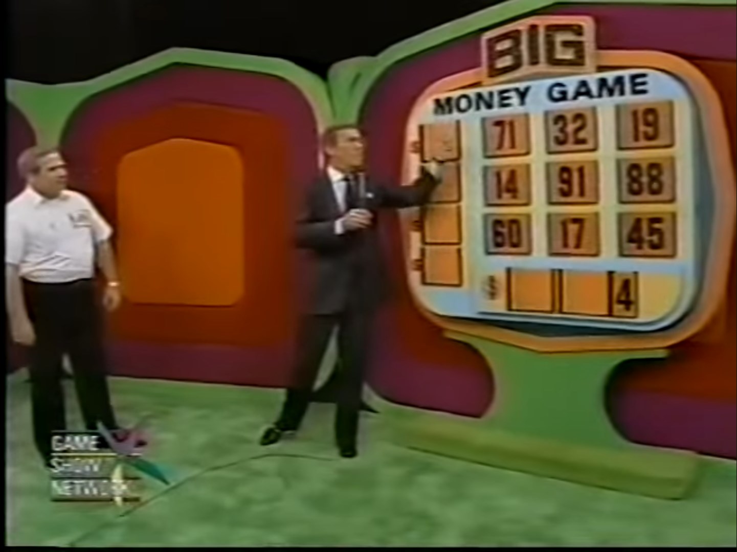 Tom Kennedy points to a board displaying the 'BIG MONEY GAME' with various numbers, while a contestant stands nearby, set in a colorful studio environment.