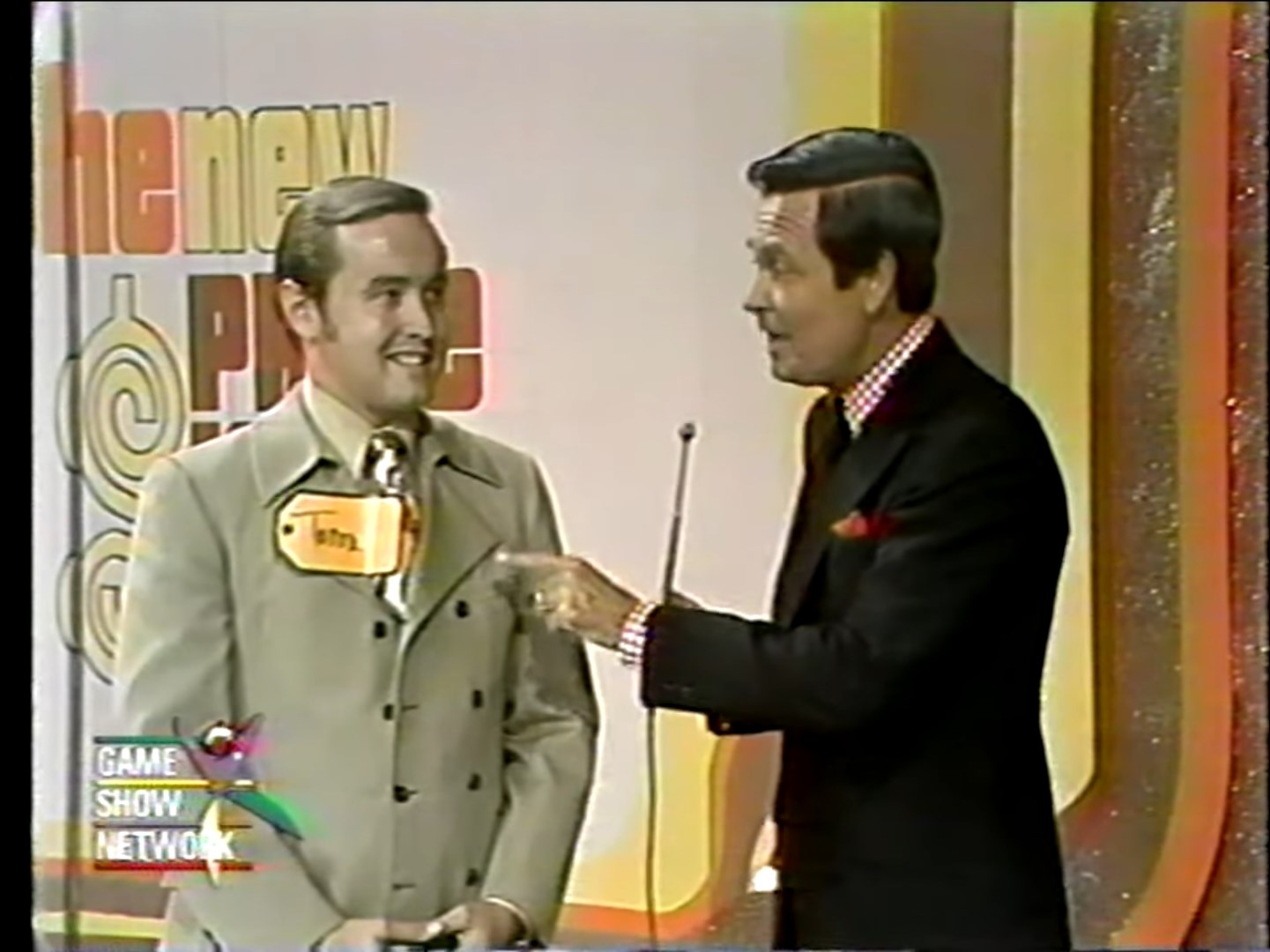 A contestant, Tom, is smiling while standing next to Bob Barker, who is explaining the rules of the game on a colorful set of The New Price is Right in 1972.