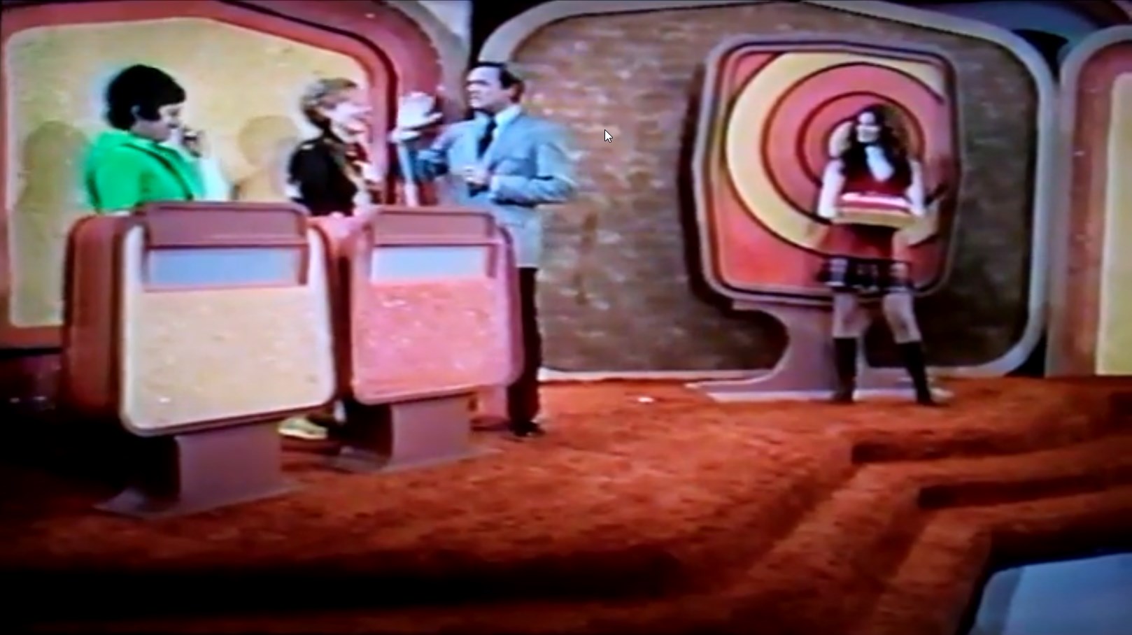 Contestants stand behind podiums during the Double Bullseye segment of game show The Price is Right, with a host engaging with one of them and a model displaying a prize in the background.