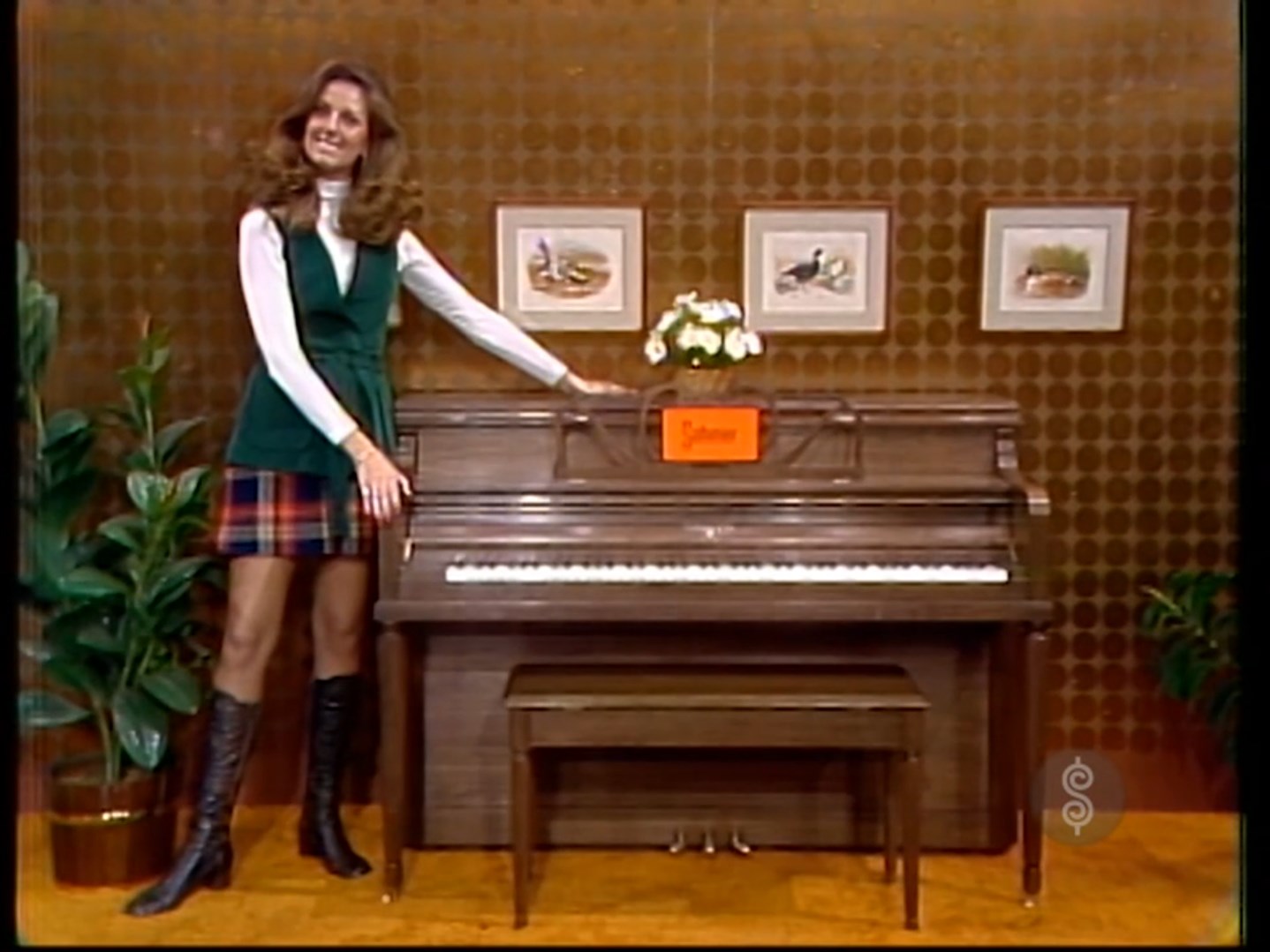 A woman stands beside a piano and bench, showcasing them as a prize on a game show set, with decorative plants and artwork in the background.