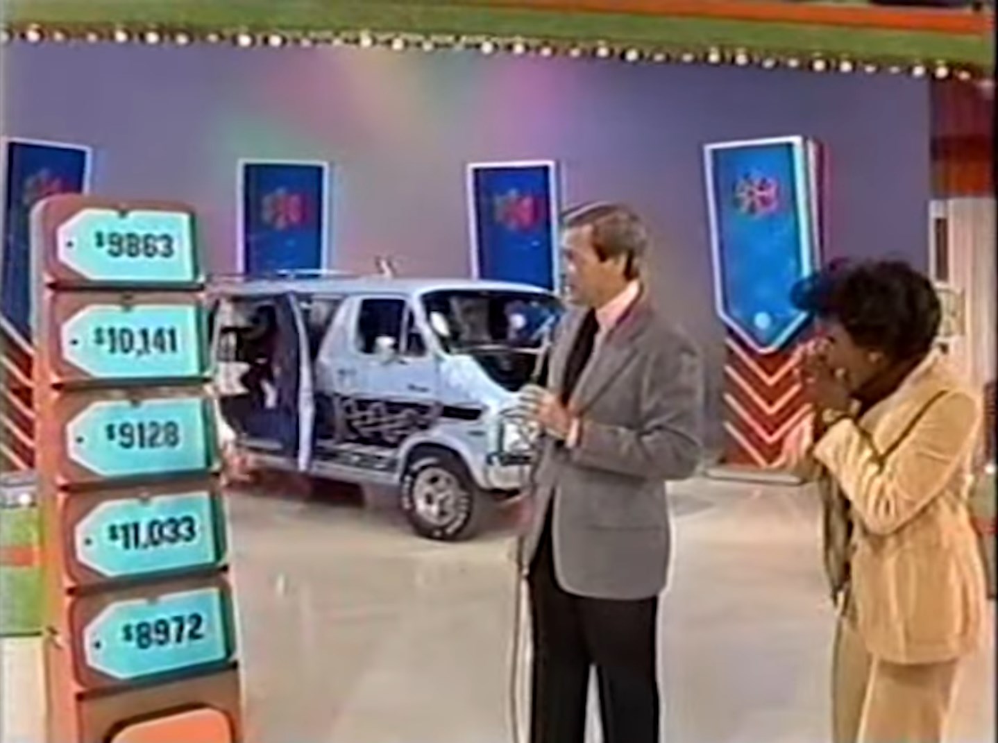 A game show host stands next to a contestant in front of a display featuring five price tags. A vehicle is partially visible in the background.