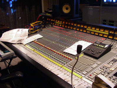 A close-up view of a recording studio mixing console with various colored knobs, faders, and papers on the surface.