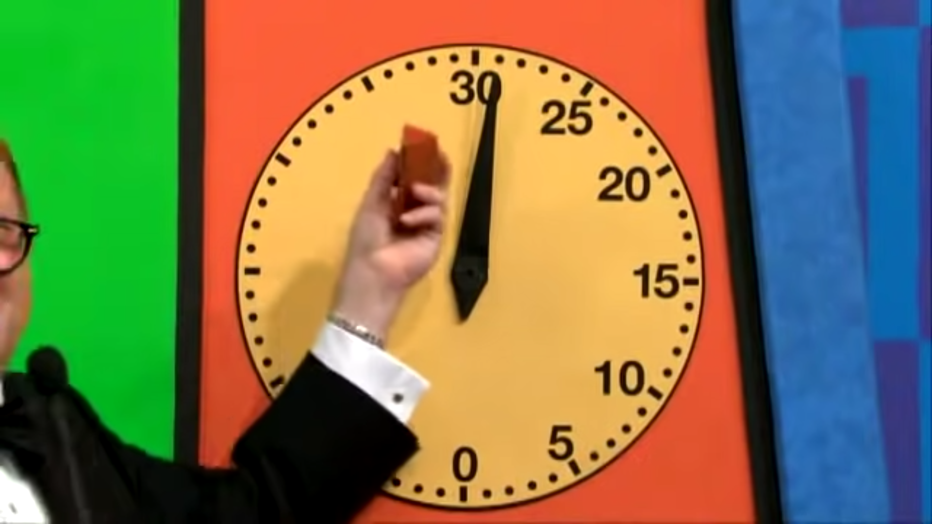 A host is holding a red object near a large yellow clock displaying the time at 30 seconds, set against a colorful background.