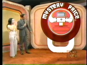 Bob Barker and a female contestant stand beside a large game display featuring the words 'MYSTERY PRICE' with a question mark and a prize compartment.