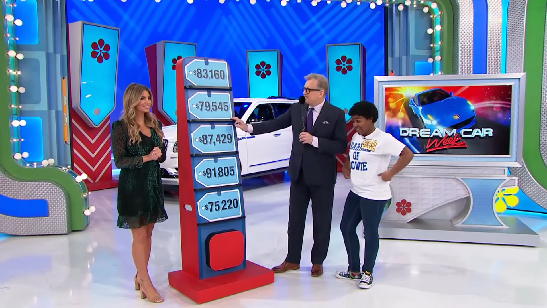 A game show set with a display of price tags on a board, featuring a host pointing to one tag while a contestant smiles nearby. A promotional graphic for 'Dream Car Week' is visible in the background, along with a white car.