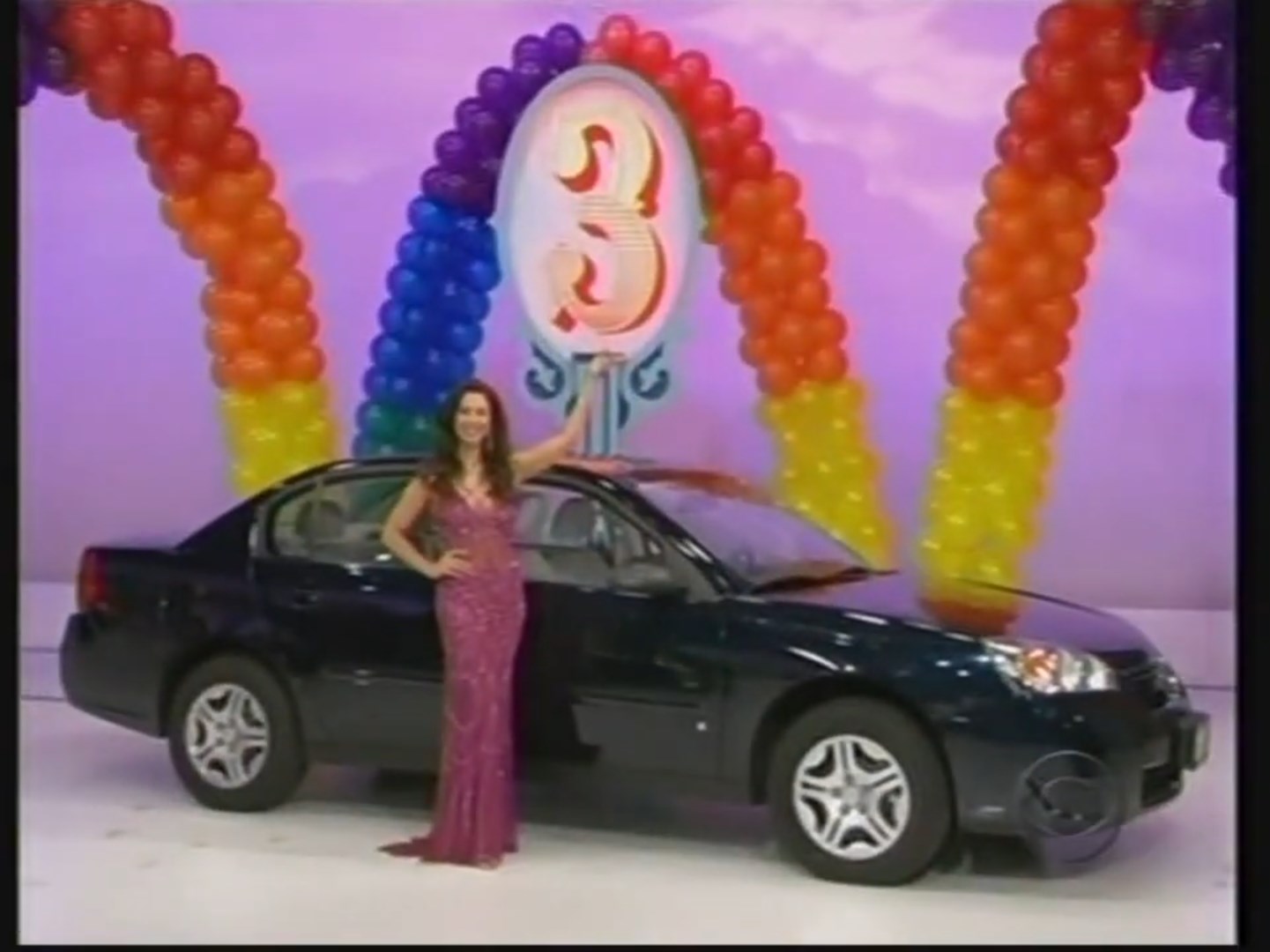 A woman in a sparkling dress stands next to a black Chevy Malibu, posing with a hand raised beside a colorful balloon arch with the number 3 displayed.