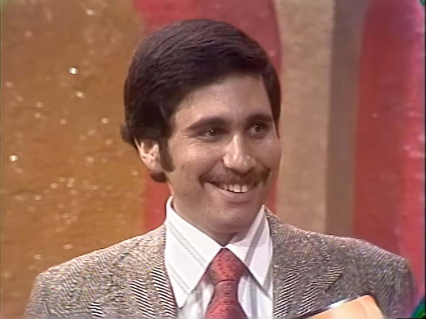 A smiling contestant with a mustache, wearing a patterned suit and red tie, stands in front of a colorful game show backdrop.