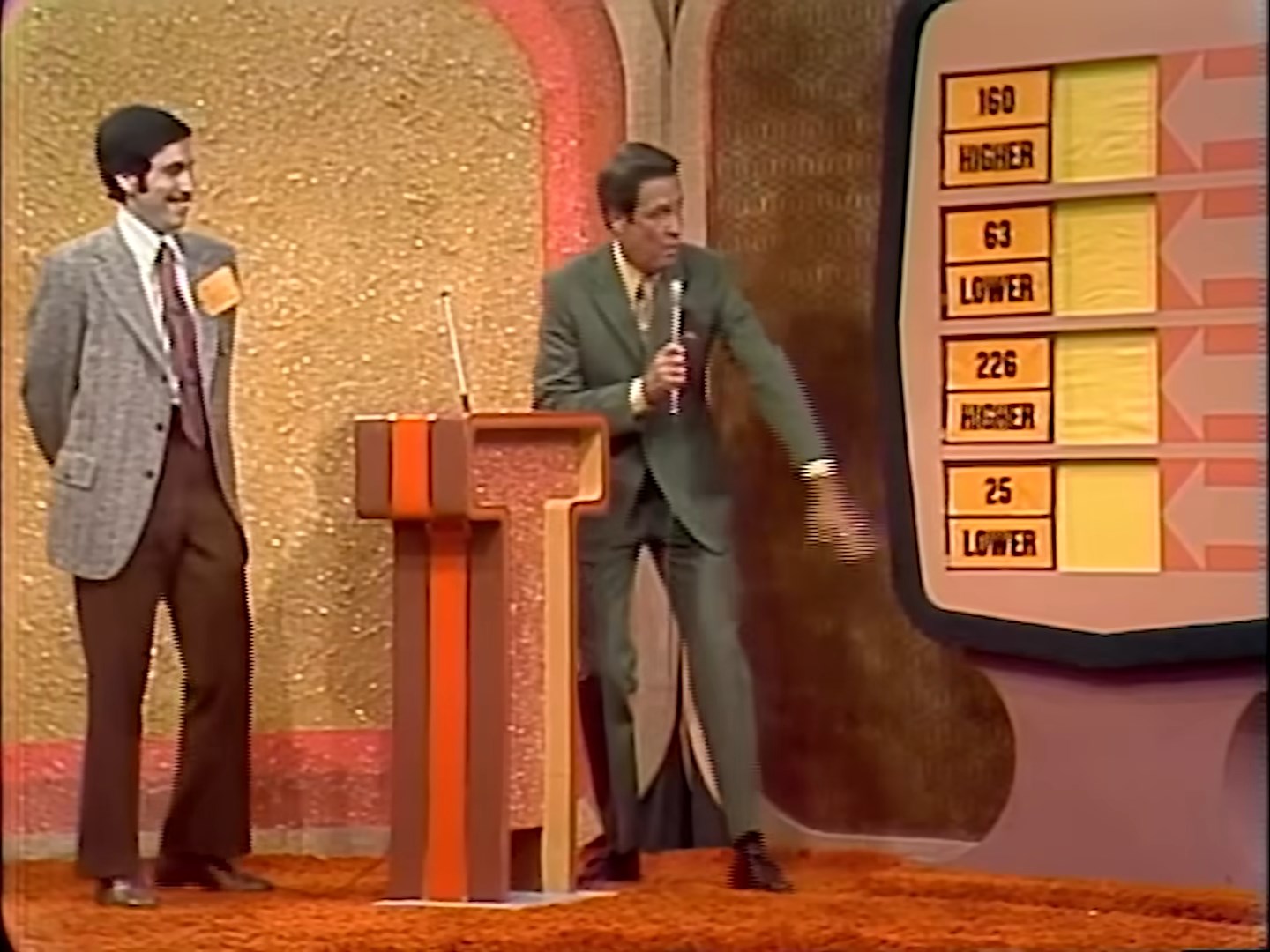 A television game show host gestures excitedly while explaining the gameplay, with a contestant smiling next to him. A large display shows numbers and indicators for higher or lower.