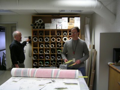 Two men are in a workspace filled with rolls of paper. One man is holding a camera while the other is standing near a table with a partially unrolled pink and white paper roll.