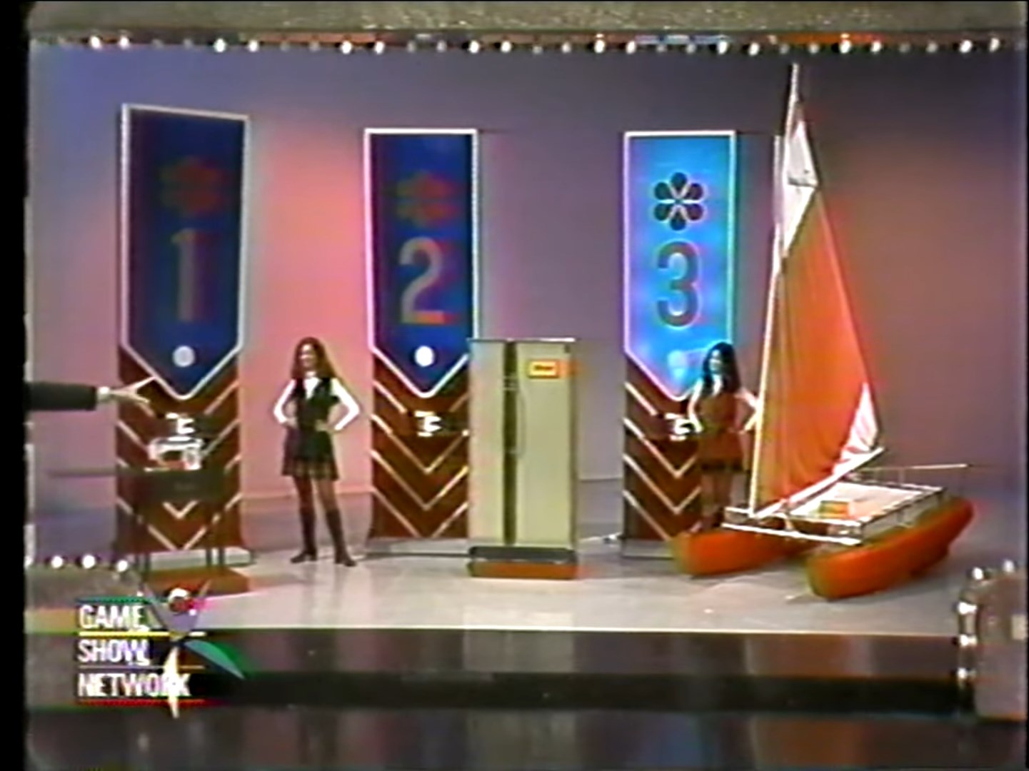 A game show set featuring three prize displays labeled 1, 2, and 3. Contestants can choose between a sewing machine, a refrigerator/freezer, and a catamaran, with a host pointing towards the prizes.