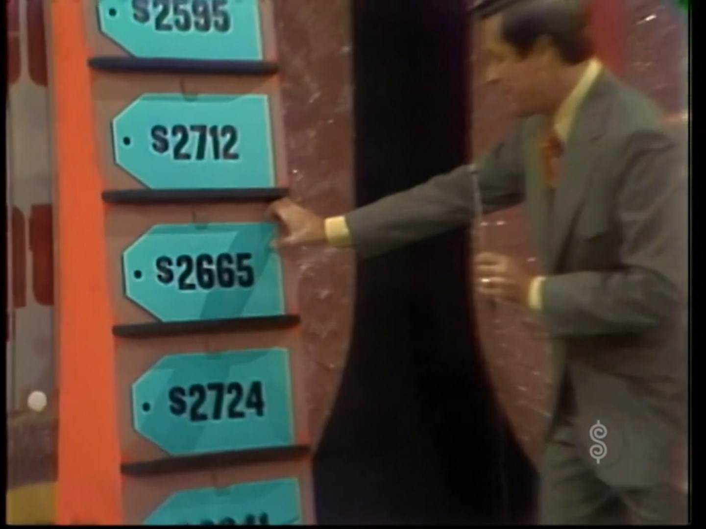 A game show host points at price tags on a display board featuring prices ranging from 95 to 12, with one highlighted price of 65.