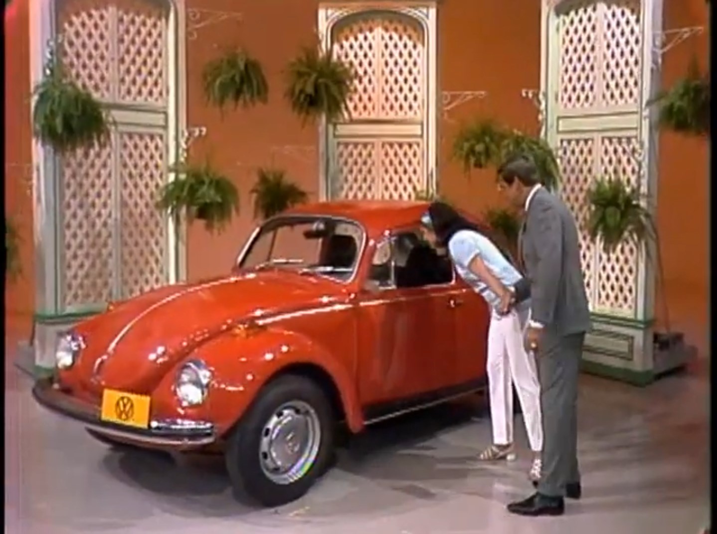 A contestant and host admire a red 1972 Volkswagen Beetle on a stage decorated with plants and privacy screens.