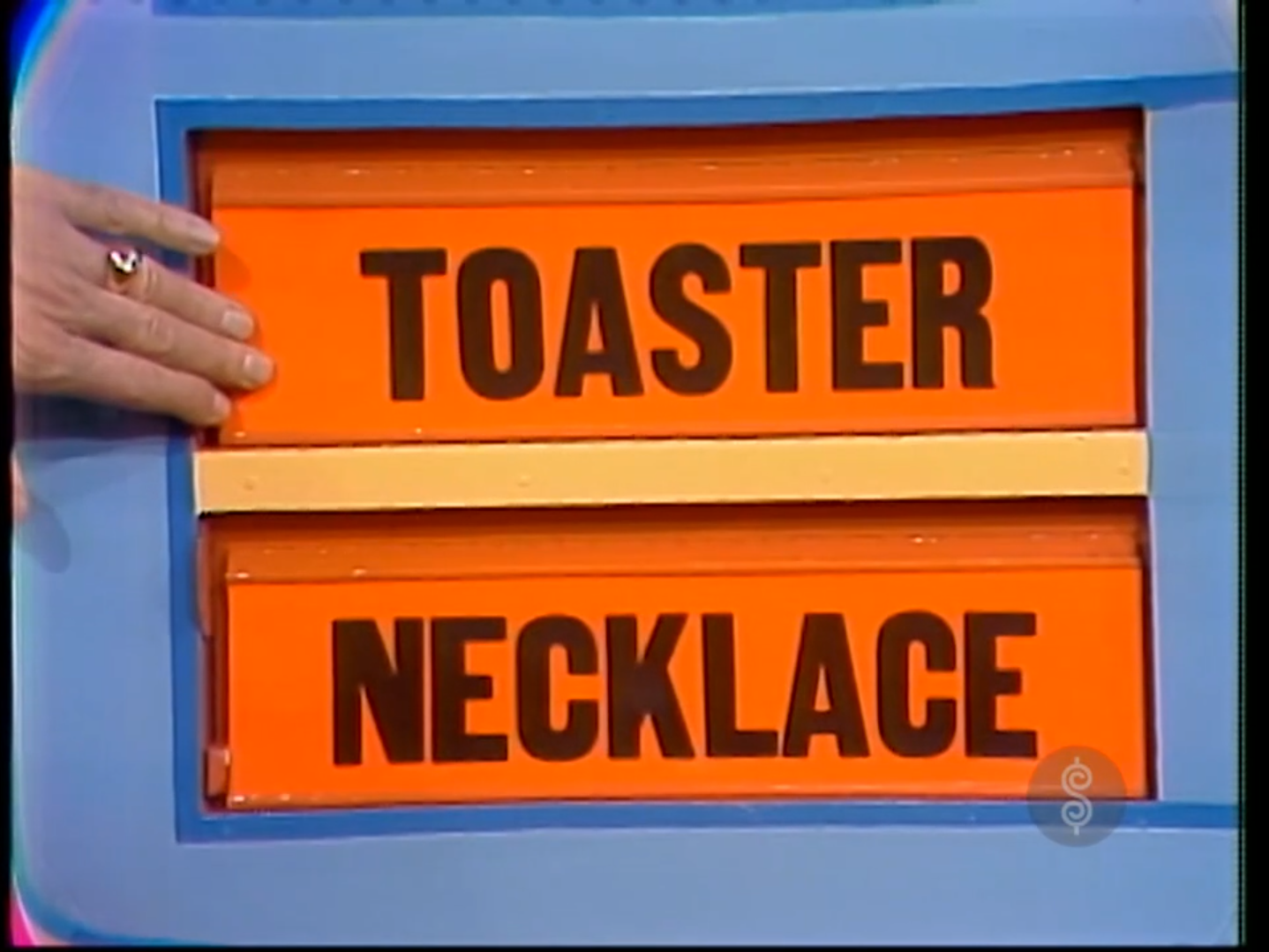 A game board displaying the options 'TOASTER' on top and 'NECKLACE' below, with a hand hovering over the board.