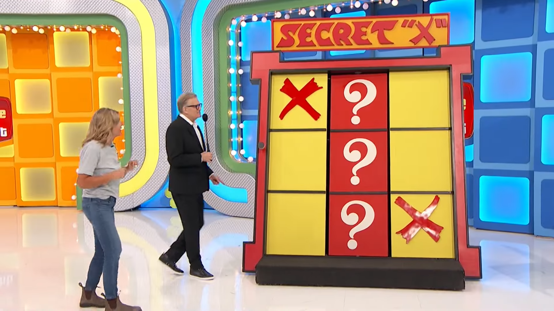 A contestant stands near the Secret 'X' game board on The Price is Right, displaying a Tic-Tac-Toe grid. The middle column is blocked, and two squares show 'X's while the remaining squares are marked with question marks.