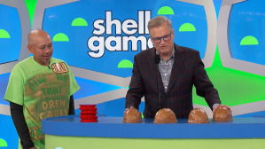 A contestant and a host participate in the 'Shell Game' on The Price is Right, with four shell-like cups and a backdrop featuring the game's name.