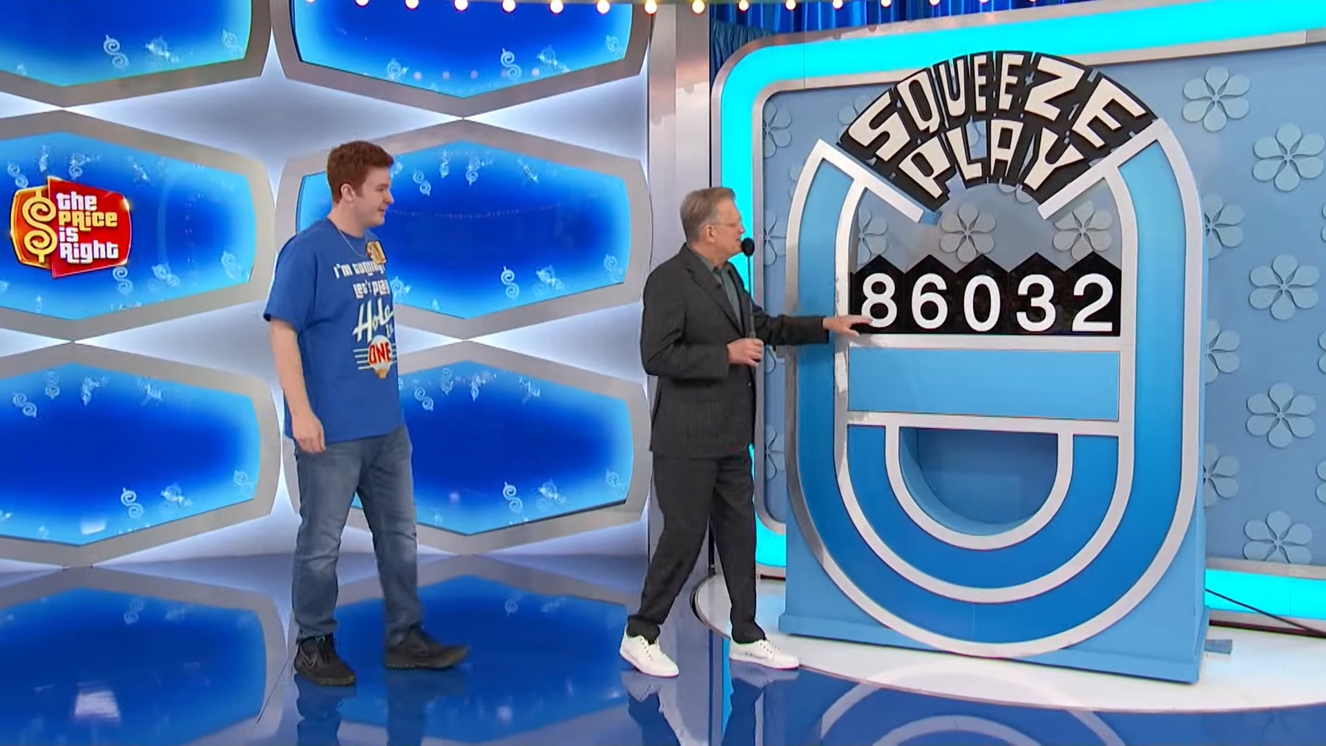 A contestant smiles next to Drew Carey, who points to the Squeeze Play game board displaying the number sequence 86032.
