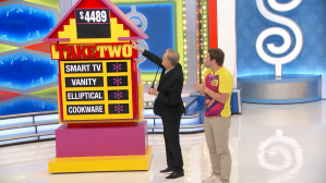 Drew Carey points to a colorful game board labeled 'TAKE TWO', with a contestant standing beside them, in a vibrant studio setting.
