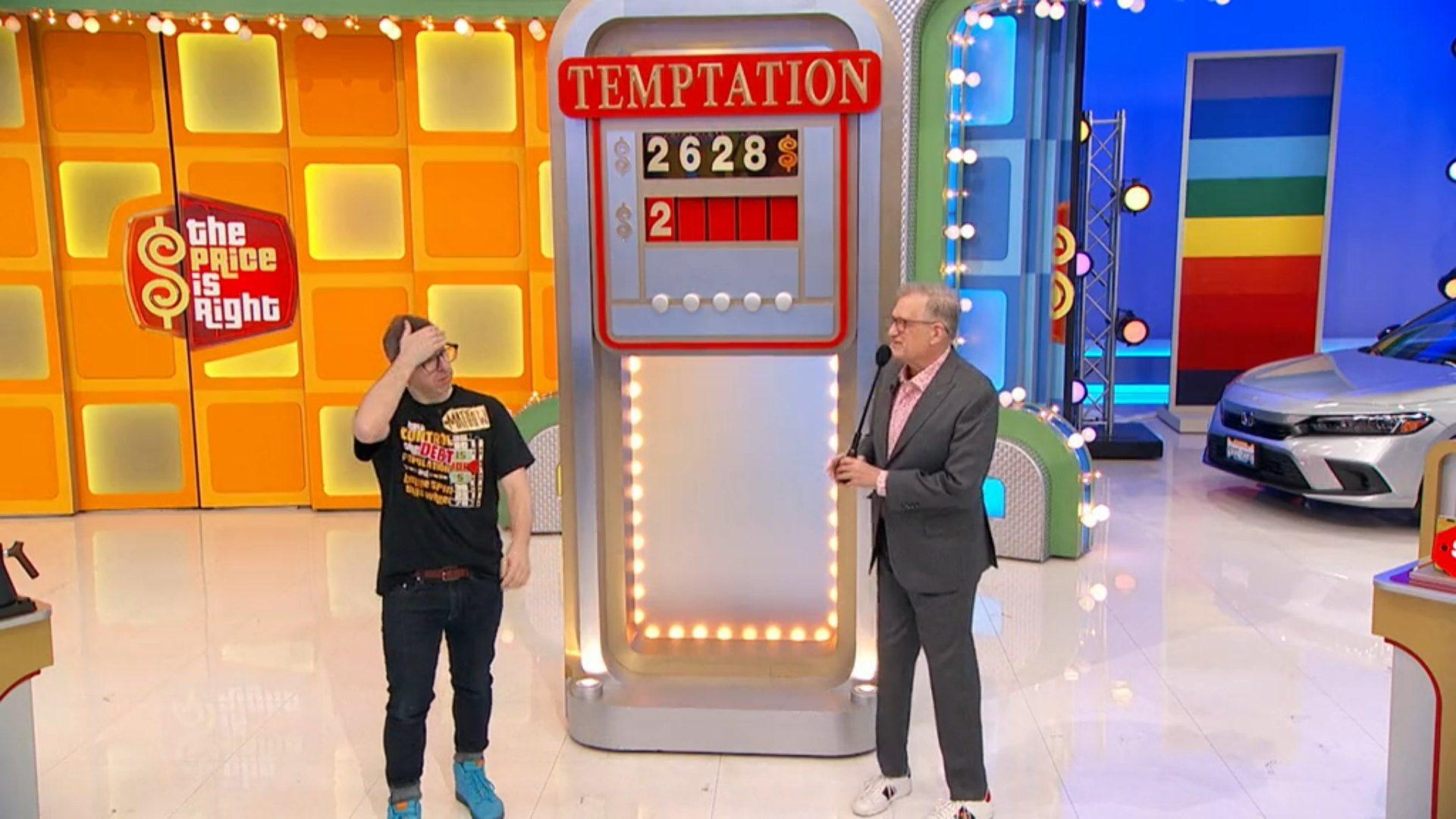 A scene from the game show 'The Price is Right', featuring the game 'Temptation'. The contestant appears concerned while standing next to the host, with a game board displaying the numbers 2628 and a blank space. The colorful set includes the show's logo in the background.